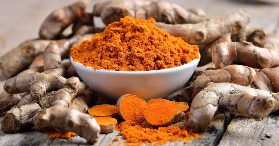 Bowl of ground turmeric powder surrounded by fresh turmeric roots on a rustic wood table
