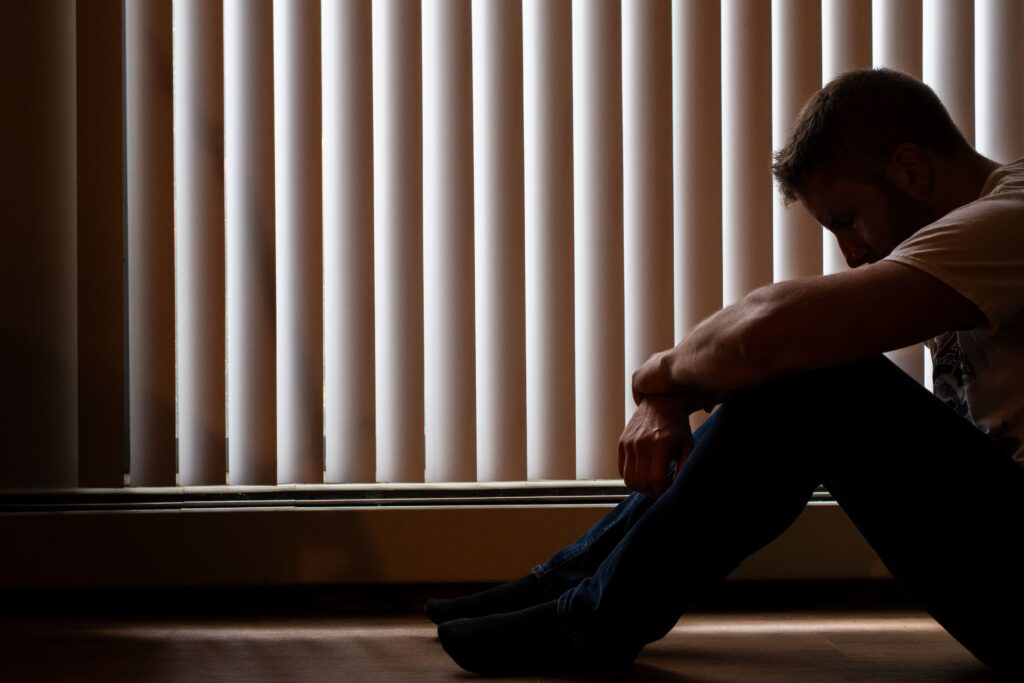 Man sitting on the floor with his head down beside window blinds, showing depression and anxiety linked to low testosterone