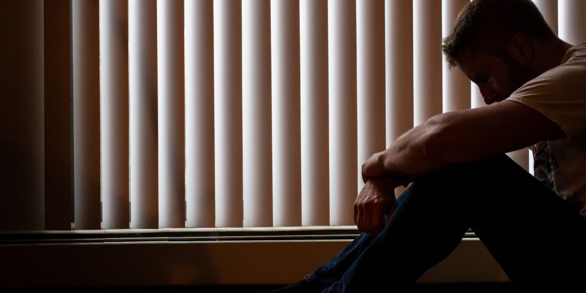 Man sitting on the floor with his head down beside window blinds, showing depression and anxiety linked to low testosterone